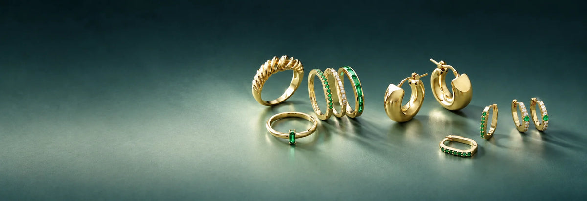 Yellow gold rings with green stones and small hoop earrings on a teal background